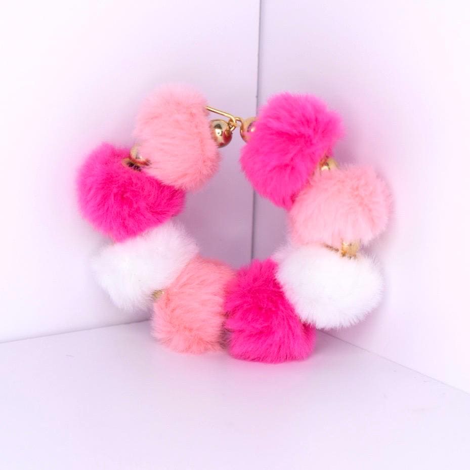 A colorful pom-pom bracelet featuring a mix of pink, white, and fuchsia pom-poms. The bracelet is connected with gold-colored metal and small bells. The fluffy texture and bright colors make it a fun accessory.