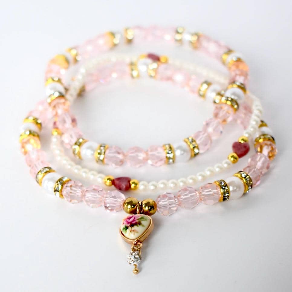 Three beaded bracelets in shades of pink and white. The bracelets feature a mix of pink faceted beads, white pearl-like beads, and gold-colored accents. One bracelet has a heart-shaped pendant with a floral design and a dangling crystal.
