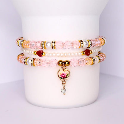 Vintage rose tumbler bracelet set with 3 beaded cup bands designed for 40 oz tumblers like Stanley features soft pink crystal beads pearls rhinestone spacers and a gold heart charm with a painted rose and dangling crystal stacked on a white tumbler for display romantic and elegant drinkware accessory perfect for bridal gifts tea lovers and vintage-inspired aesthetics