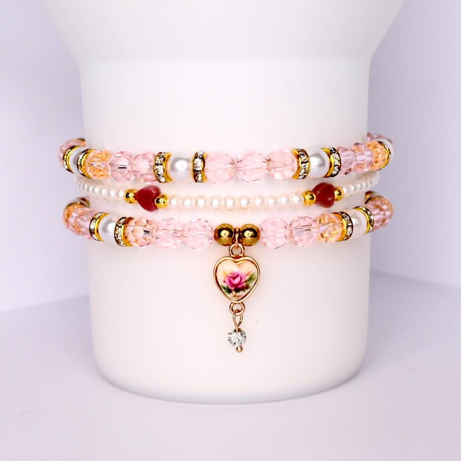 Vintage rose tumbler bracelet set with 3 beaded cup bands designed for 40 oz tumblers like Stanley features soft pink crystal beads pearls rhinestone spacers and a gold heart charm with a painted rose and dangling crystal stacked on a white tumbler for display romantic and elegant drinkware accessory perfect for bridal gifts tea lovers and vintage-inspired aesthetics