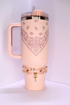 A pink conch floral Stanley tumbler with a handle, a lid, and a straw. The tumbler has a heart-shaped floral design. Two bracelets with pink and gold beads and a heart charm are around the base of the tumbler. The background is a light pink color.