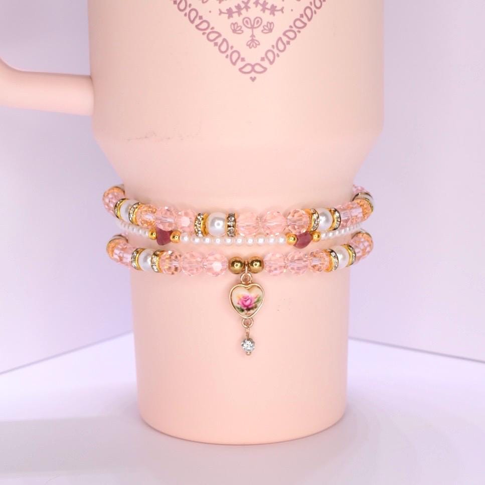 A pink mug adorned with two beaded bracelets. The bracelets feature pink, gold, and pearl beads. The lower bracelet has a heart-shaped charm with a rose design and a dangling crystal. The mug has a handle and a decorative pattern near the top.