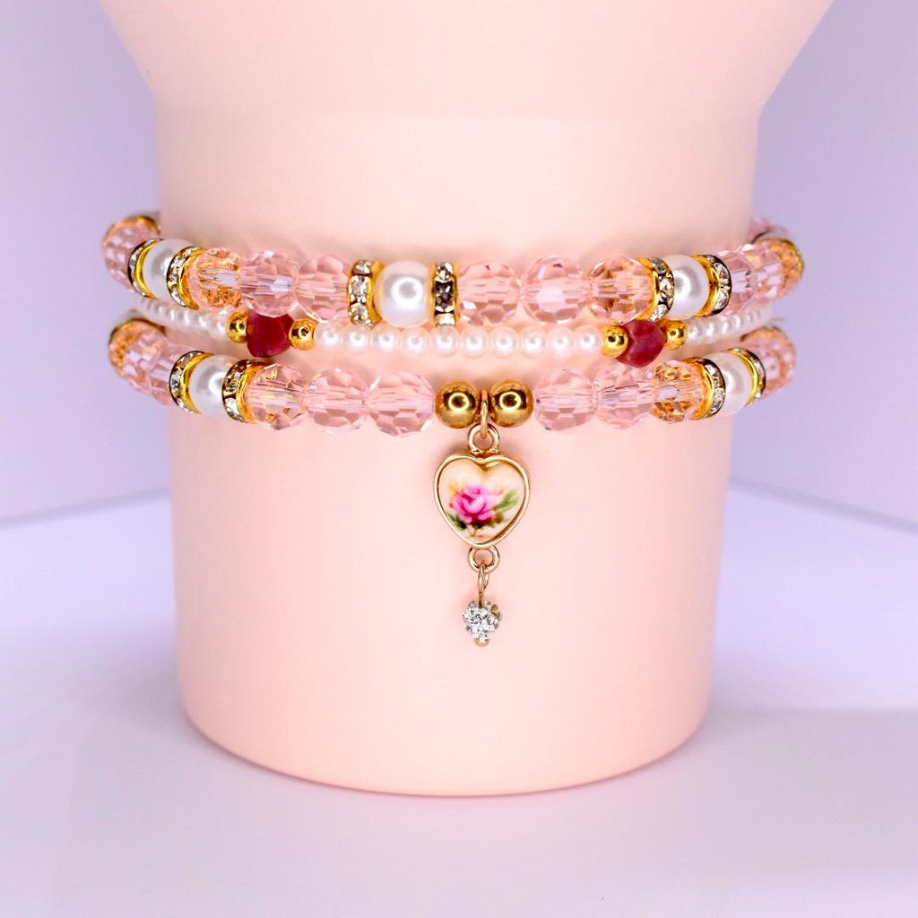 Three pink beaded bracelets with gold accents and a heart charm. The bracelets feature pink beads, pearls, and gold-colored spacers. The heart charm has a pink rose design and a small dangling crystal.
