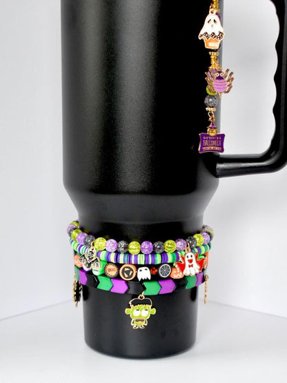 A black tumbler adorned with Halloween-themed accessories. A charm hangs from the handle, featuring a cupcake, owl, and sign that reads 'Happy Halloween.' Around the base are stacked bracelets with beads in green, purple, and black, along with Halloween charms like a ghost and Frankenstein's monster.