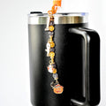 A black travel mug with a silver rim and handle, adorned with a Halloween-themed charm. The charm features a series of beads and pendants, including a ghost, a pumpkin, and other festive elements.