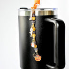 A black travel mug with a silver rim and handle, adorned with a Halloween-themed charm. The charm features a series of beads and pendants, including a ghost, a pumpkin, and other festive elements.