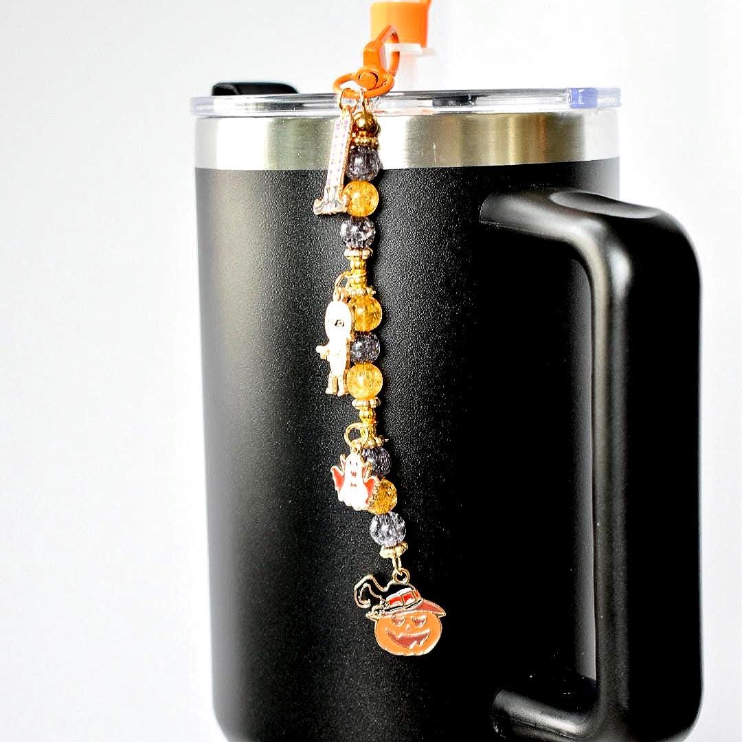 A black travel mug with a silver rim and handle, adorned with a Halloween-themed charm. The charm features a series of beads and pendants, including a ghost, a pumpkin, and other festive elements.