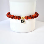 A basketball-themed bracelet featuring orange beads with black lines, resembling basketballs. The bracelet includes a gold charm with the letter "K" on a black background. The bracelet is displayed on a white surface.