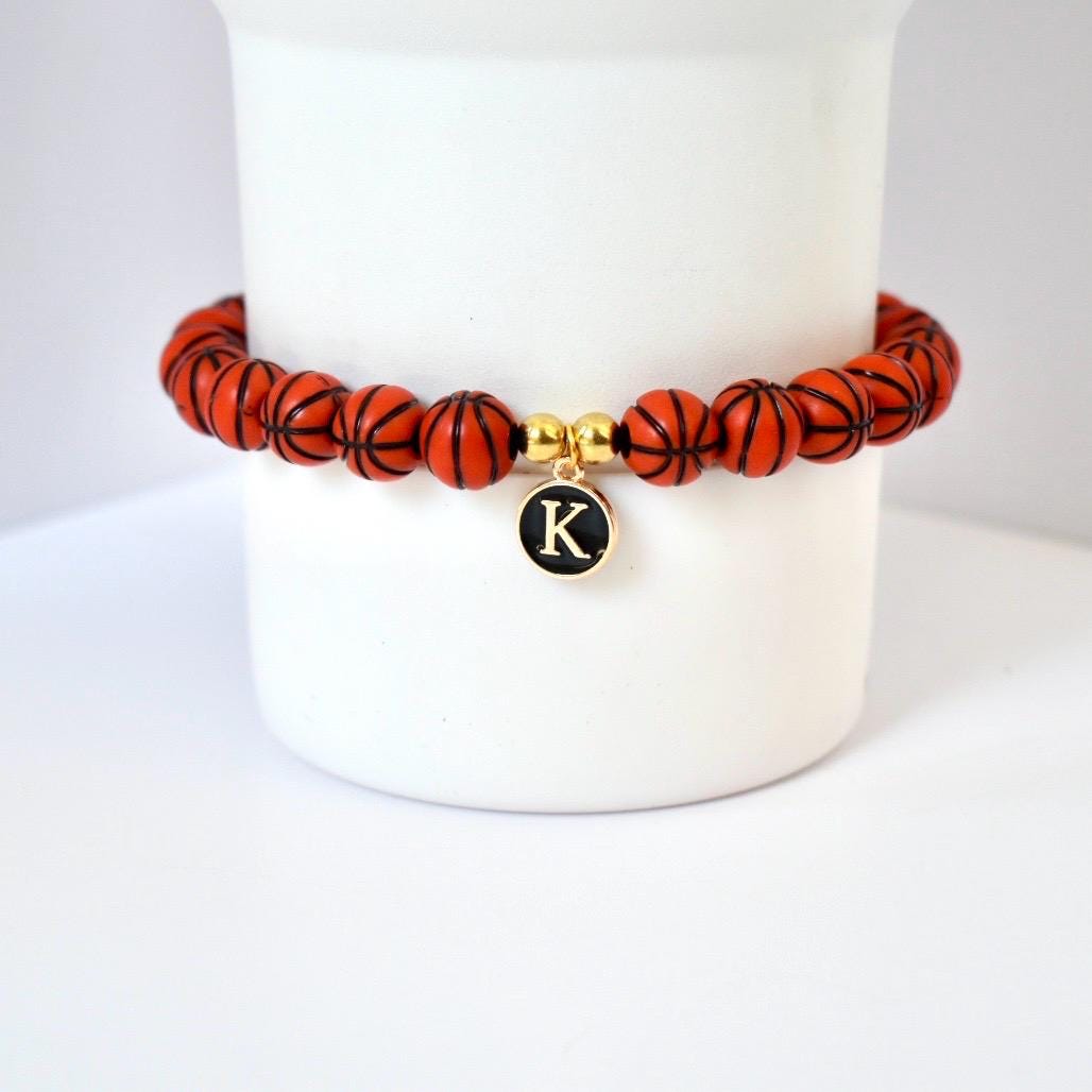 A basketball-themed bracelet featuring orange beads with black lines, resembling basketballs. The bracelet includes a gold charm with the letter "K" on a black background. The bracelet is displayed on a white surface.