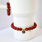 A basketball-themed bracelet and a matching stack of beads. The bracelet features orange beads with black lines, resembling basketballs. A gold charm with a black 'K' hangs from the bracelet. The beads are displayed on a white mug.