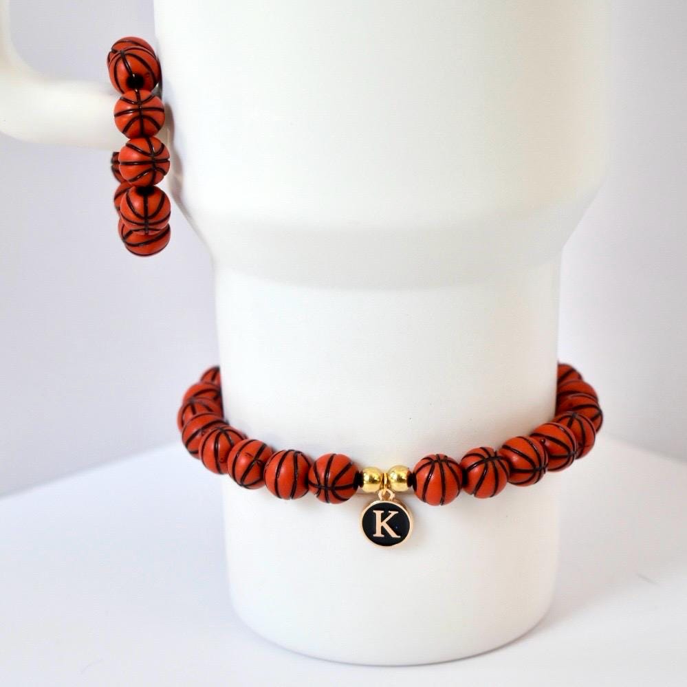 A basketball-themed bracelet and a matching stack of beads. The bracelet features orange beads with black lines, resembling basketballs. A gold charm with a black 'K' hangs from the bracelet. The beads are displayed on a white mug.