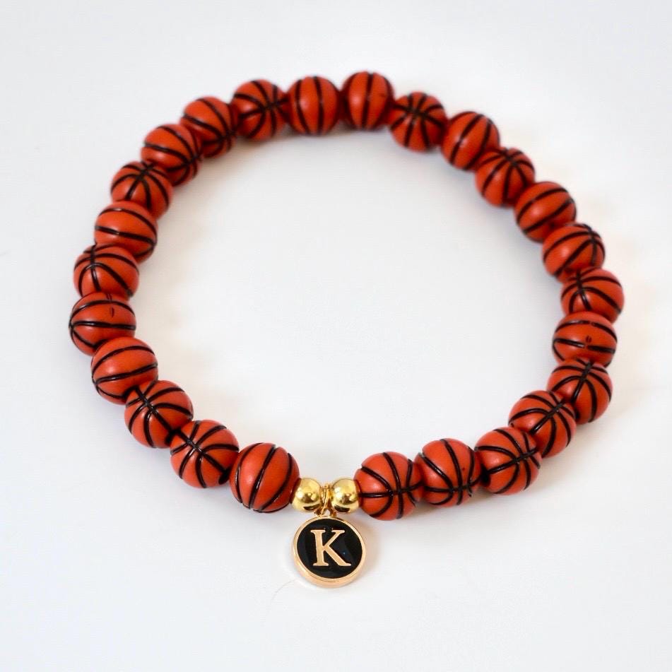 A beaded bracelet featuring orange basketball-shaped beads with black lines. A gold charm with the letter "K" in black hangs from the bracelet. The bracelet is on a white background.