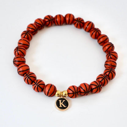 A beaded bracelet featuring orange basketball-shaped beads with black lines. A gold charm with the letter "K" in black hangs from the bracelet. The bracelet is on a white background.