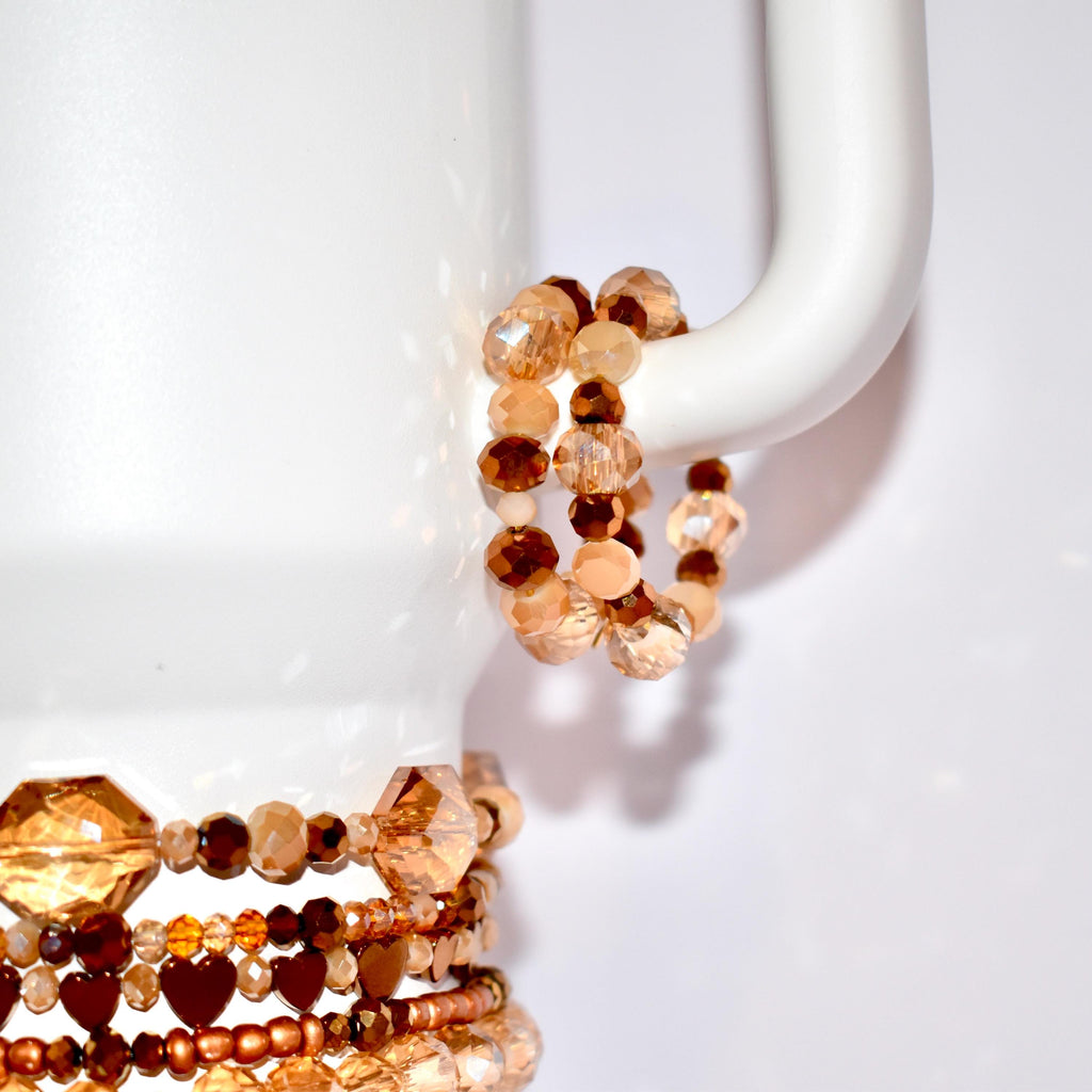 A collection of beaded bracelets in shades of brown, tan, and peach. The bracelets feature various bead shapes, including faceted and round beads. Some bracelets have heart-shaped beads. The bracelets are displayed on a white surface.