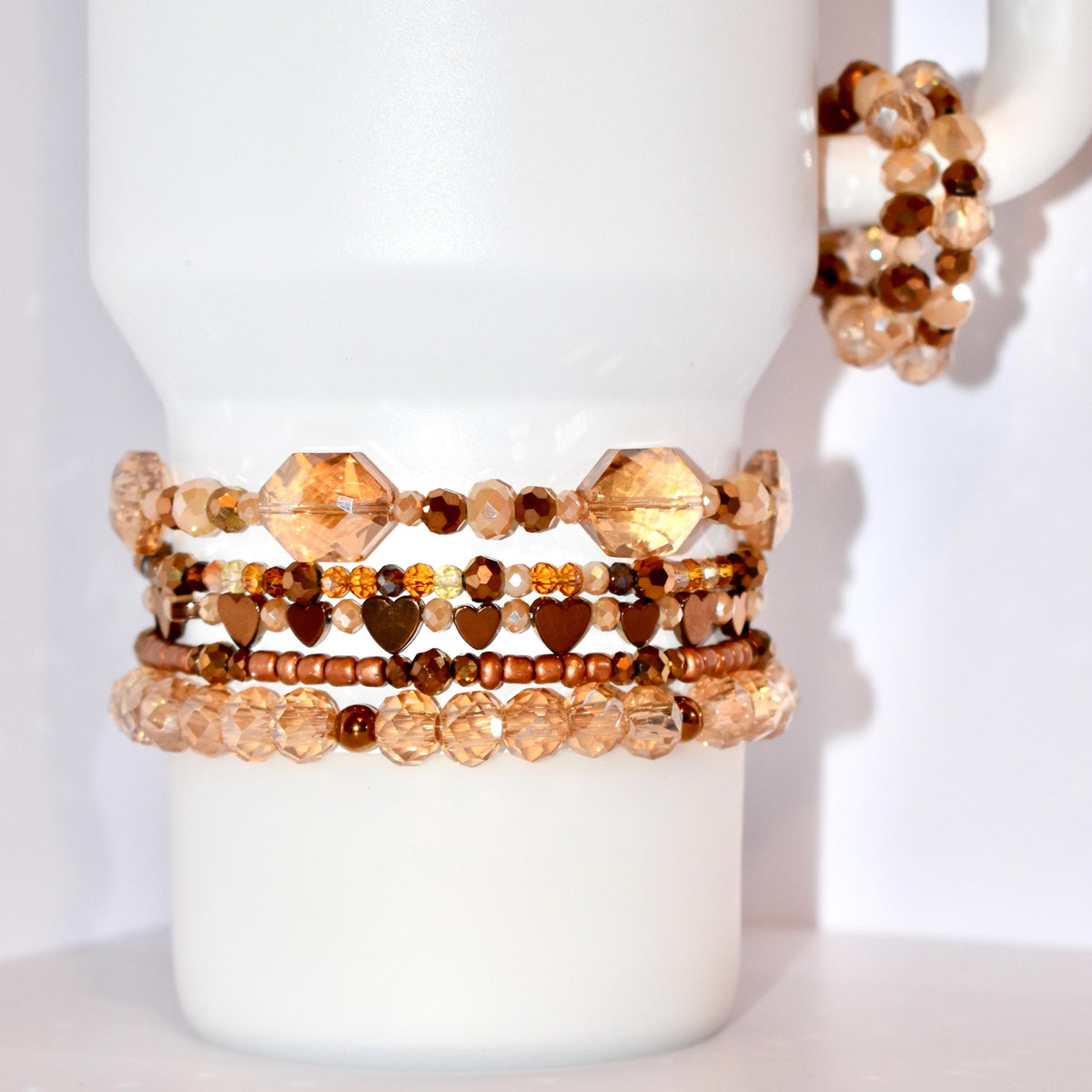 A collection of beaded bracelets in shades of brown and gold. The bracelets feature various bead shapes, including faceted crystals, small round beads, and heart-shaped beads. One bracelet is wrapped around a white mug handle.