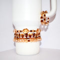 A collection of beaded bracelets in shades of brown and gold. The bracelets feature various bead shapes, including faceted beads and heart-shaped beads. Some bracelets are stacked together, while others are displayed separately.