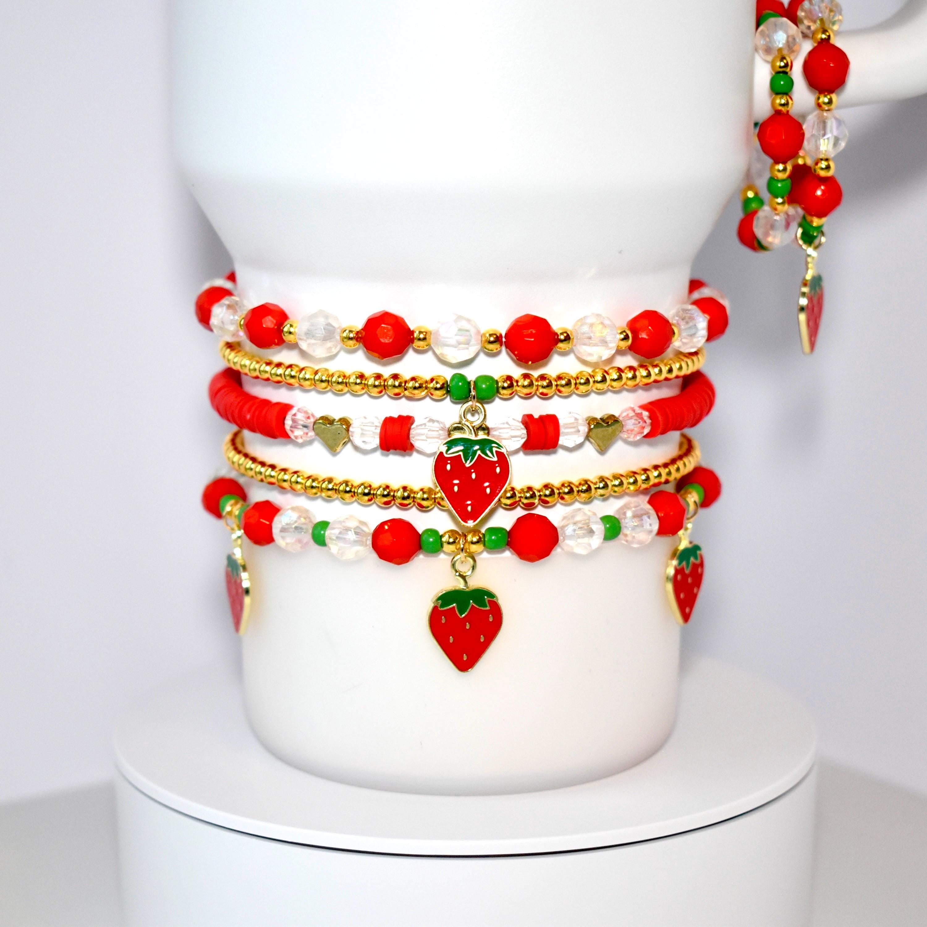 A stack of five beaded tumbler bracelets around the base of a white Stanley tumbler. Two of the bracelets are simple gold plastic beads. The other three are a mix of red, green, and clear beads with dangling strawberry charms.