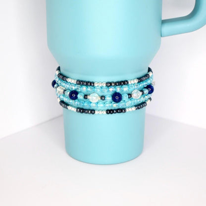 Stack of five handmade beaded tumbler bracelets in shades of blue, silver, and black on a blue tumbler, featuring glass and pearl-like beads.