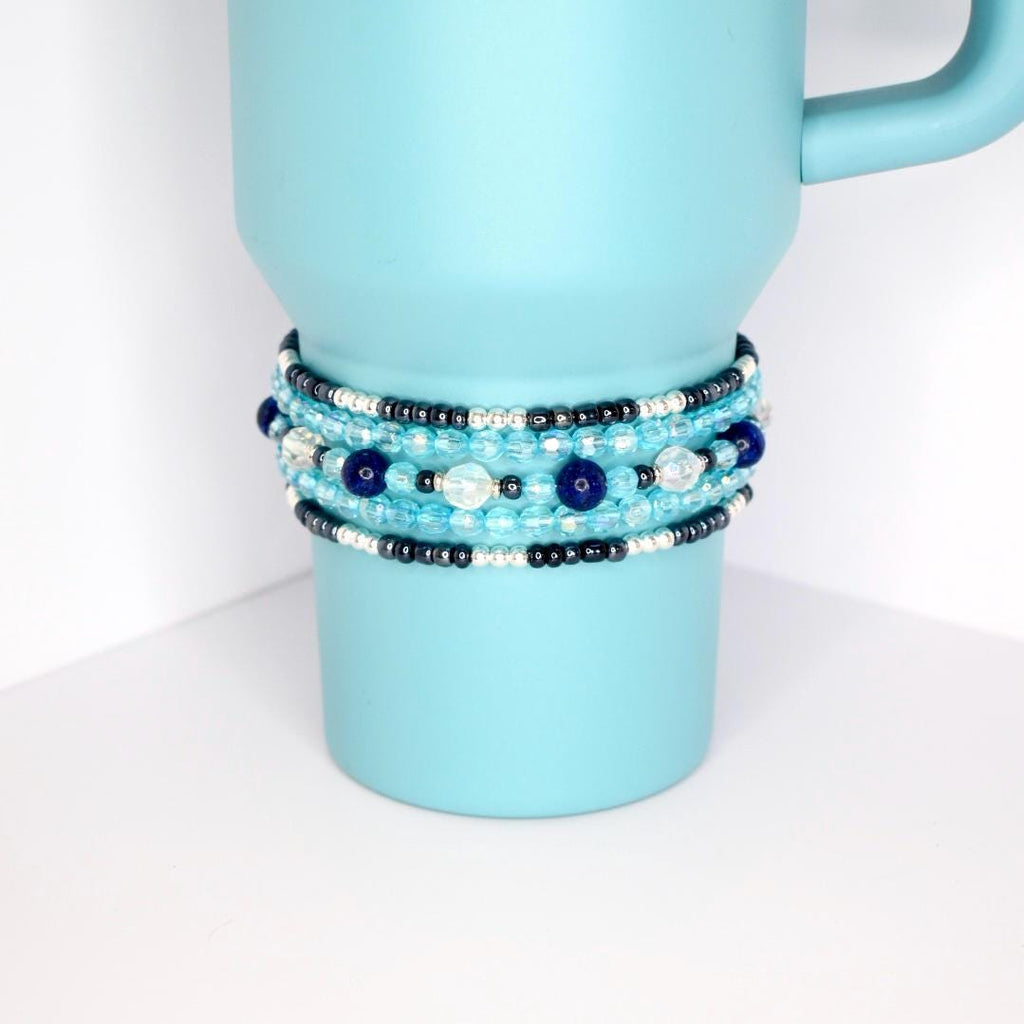 Stack of five handmade beaded tumbler bracelets in shades of blue, silver, and black on a blue tumbler, featuring glass and pearl-like beads.