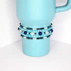 Stack of five handmade beaded tumbler bracelets in shades of blue, silver, and black on a blue tumbler, featuring glass and pearl-like beads.