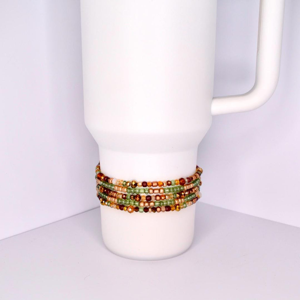 A white tumbler with a beaded bracelet wrapped around it. The bracelet features multiple strands of small, colorful beads in shades of green, gold, brown, and red. The beads are tightly strung together, creating a textured band around the tumbler.