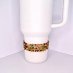 A white tumbler with a beaded bracelet wrapped around it. The bracelet features multiple strands of small, colorful beads in shades of green, gold, brown, and red. The beads are tightly strung together, creating a textured band around the tumbler.