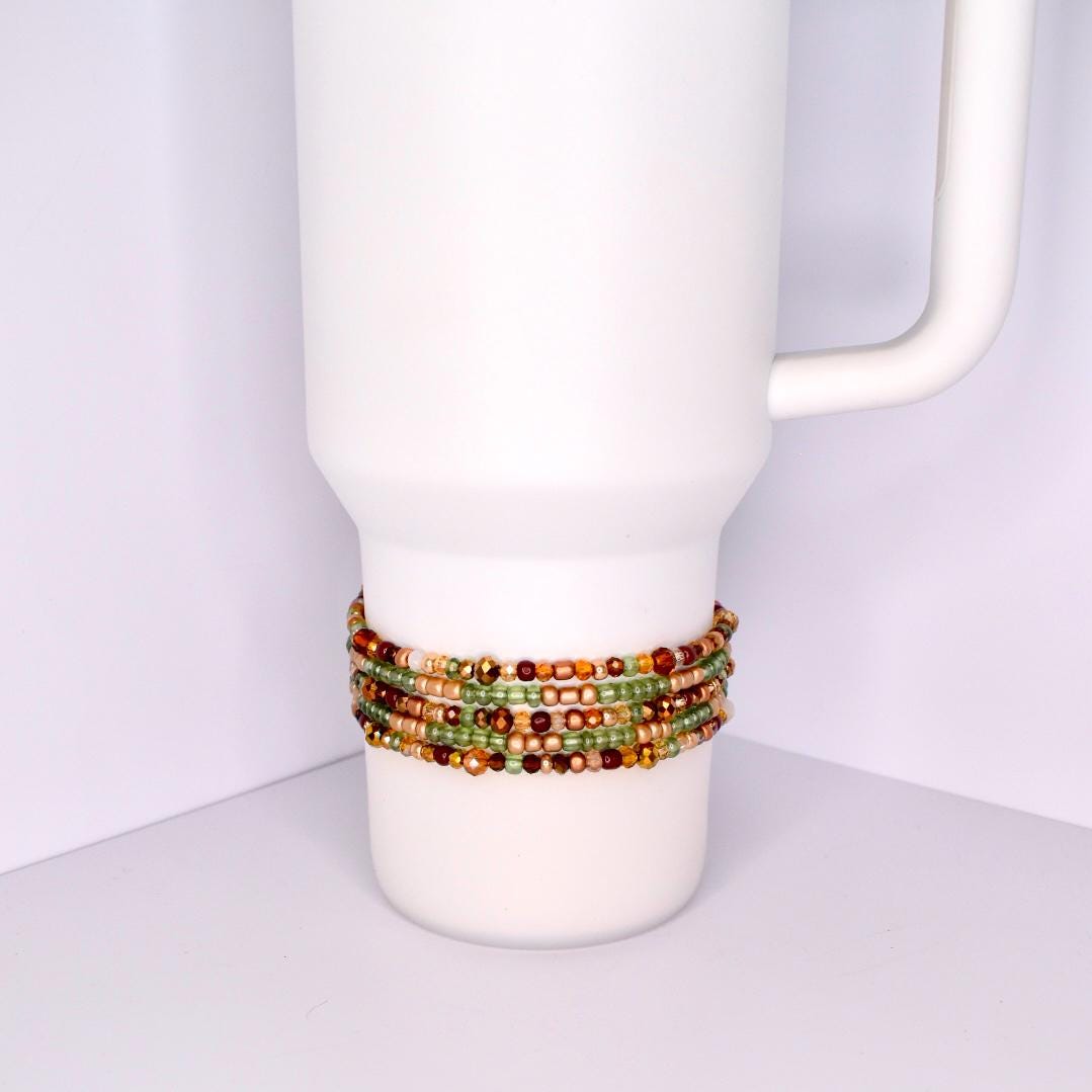 A white tumbler with a beaded bracelet wrapped around it. The bracelet features multiple strands of small, colorful beads in shades of green, gold, brown, and red. The beads are tightly strung together, creating a textured band around the tumbler.