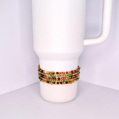 A white tumbler with a beaded bracelet wrapped around it. The bracelet features multiple strands of small, colorful beads in shades of green, gold, brown, and red. The beads are tightly strung together, creating a textured band around the tumbler.