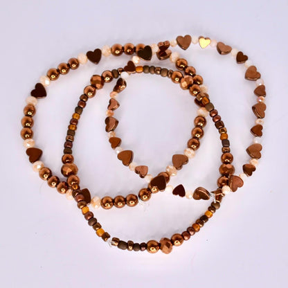 Three beaded bracelets in shades of brown and gold. The bracelets feature a mix of small round beads, heart-shaped beads, and crystal-like beads. The beads are strung together to create a layered effect, with the bracelets overlapping each other. The overall aesthetic is delicate and feminine.
