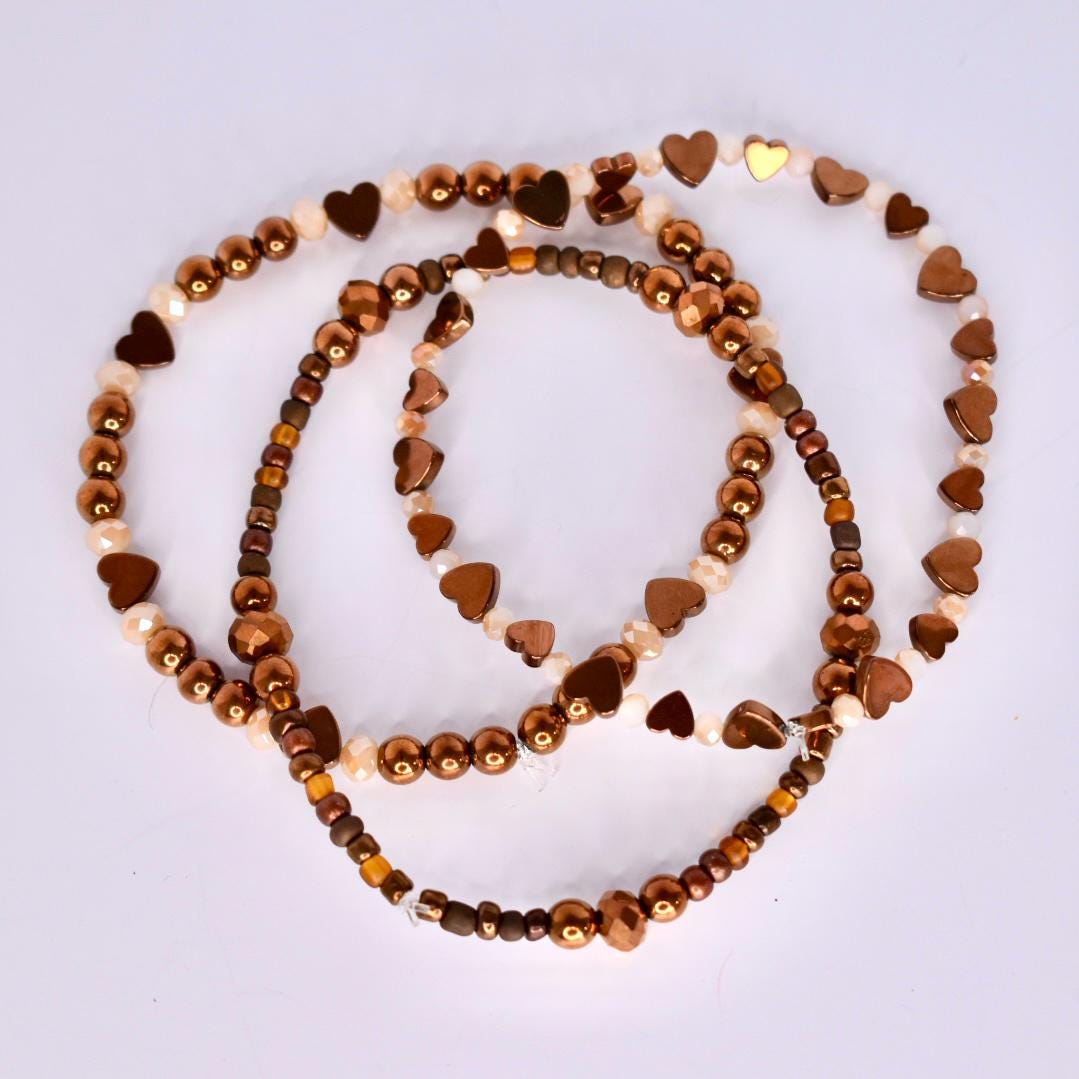 Three beaded bracelets in shades of brown and gold. The bracelets feature a mix of small round beads, heart-shaped beads, and crystal-like beads. The beads are strung together to create a layered effect, with the bracelets overlapping each other. The overall aesthetic is delicate and feminine.