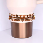 A light pink and copper-colored tumbler with three beaded bracelets stacked around the middle. The bracelets feature heart-shaped beads in shades of brown and gold, interspersed with smaller beads. The tumbler has a pink top and a metallic copper base, creating a contrast in textures and colors.