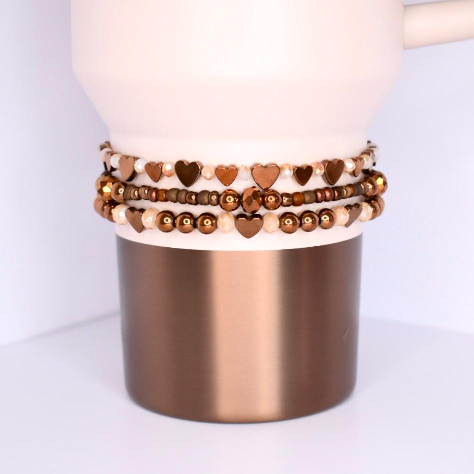 A light pink and copper-colored tumbler with three beaded bracelets stacked around the middle. The bracelets feature heart-shaped beads in shades of brown and gold, interspersed with smaller beads. The tumbler has a pink top and a metallic copper base, creating a contrast in textures and colors.