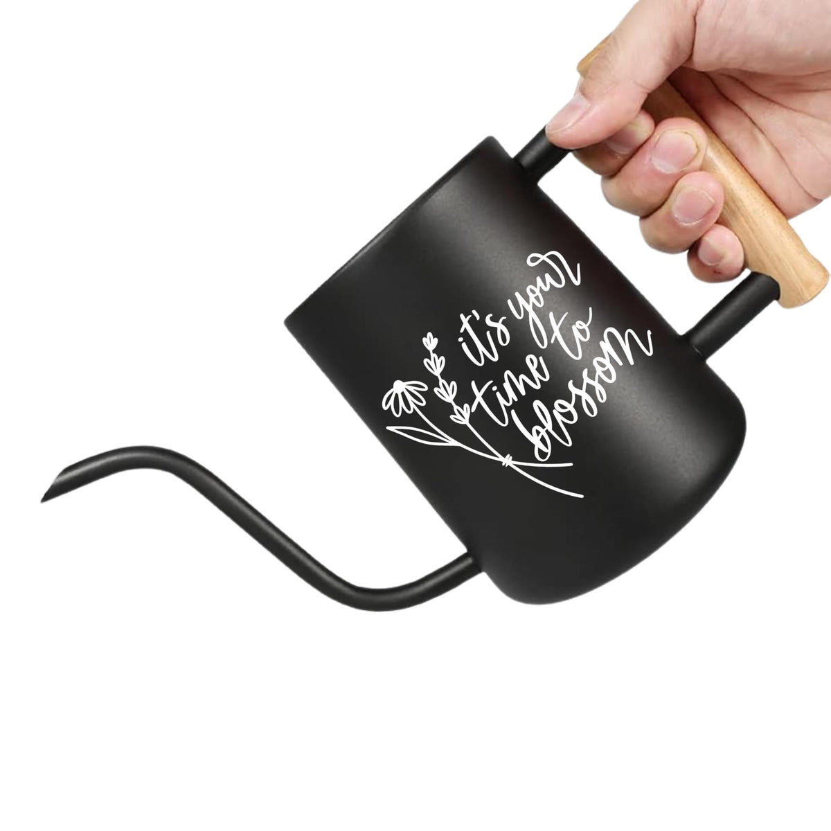 A black watering can with a wooden handle. The watering can has a long, curved spout and a black body. White script on the side reads 'it's your time to blossom' with a floral design. The can is held by a hand.