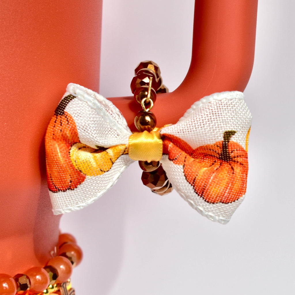 A close-up of a fall-themed tumbler handle bracelet featuring faceted bronze beads and a mini bow charm with a yellow center and pumpkin-printed ribbon, attached to the handle of a matte orange Stanley tumbler.