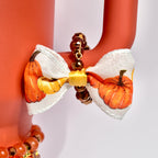 A close-up of a fall-themed tumbler handle bracelet featuring faceted bronze beads and a mini bow charm with a yellow center and pumpkin-printed ribbon, attached to the handle of a matte orange Stanley tumbler.