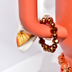 A festive tumbler charm bracelet on a rust-colored Stanley cup handle, featuring metallic brown beads and a fabric bow with pumpkins in fall shades of orange and yellow.