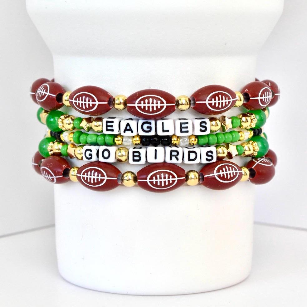 Four beaded bracelets with a football theme. The bracelets feature maroon football-shaped beads with white stitching, gold beads, green beads, and white letter beads spelling "EAGLES GO BIRDS". The bracelets are stacked on a white cylindrical object.