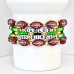 Four beaded bracelets with a football theme. The bracelets feature maroon football-shaped beads with white stitching, gold beads, green beads, and white letter beads spelling "EAGLES GO BIRDS". The bracelets are stacked on a white cylindrical object.