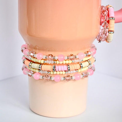 Stack of pink and pastel beaded bracelets. The bracelets are displayed on a peach-colored mug. The bracelets feature a variety of beads, including pink round beads, clear faceted beads, and flat disc beads in pastel colors.