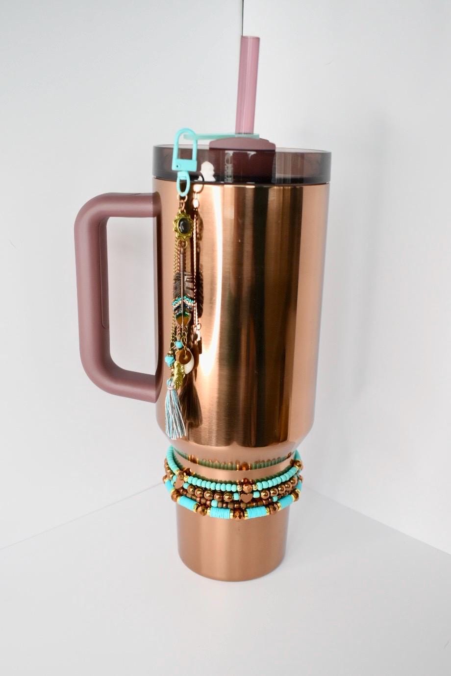 A copper-colored insulated tumbler with a handle, a lid, and a pink straw. A light blue charm with a key ring is attached to the lid. The charm has feathers, beads, and a tassel. A stack of turquoise and brown beaded bracelets is around the base of the tumbler.