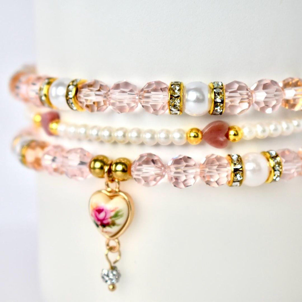 Three delicate bracelets stacked together. The top and bottom bracelets feature pink faceted beads, gold accents, and white pearl beads. The middle bracelet is made of small white pearls. A heart-shaped charm with a floral design dangles from the bottom bracelet.