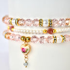 Three delicate bracelets stacked together. The top and bottom bracelets feature pink faceted beads, gold accents, and white pearl beads. The middle bracelet is made of small white pearls. A heart-shaped charm with a floral design dangles from the bottom bracelet.