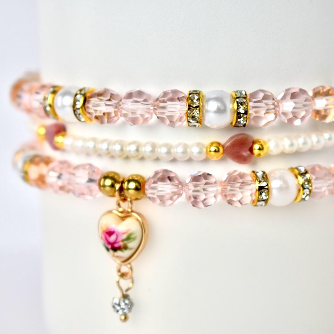 Three delicate bracelets stacked together. The top and bottom bracelets feature pink faceted beads, gold accents, and white pearl beads. The middle bracelet is made of small white pearls. A heart-shaped charm with a floral design dangles from the bottom bracelet.