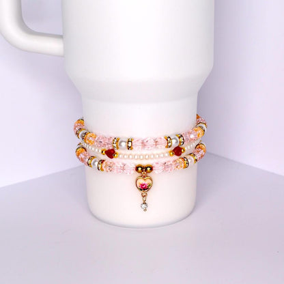 Three beaded bracelets stacked on a white cylindrical object. The bracelets feature pink, gold, and white beads. One bracelet has a heart-shaped charm with a rose design and a dangling crystal. The bracelets are adorned with gold accents and small red beads.