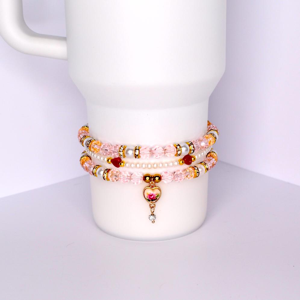 Three beaded bracelets stacked on a white cylindrical object. The bracelets feature pink, gold, and white beads. One bracelet has a heart-shaped charm with a rose design and a dangling crystal. The bracelets are adorned with gold accents and small red beads.