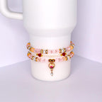 Three beaded bracelets stacked on a white cylindrical object. The bracelets feature pink, gold, and white beads. One bracelet has a heart-shaped charm with a rose design and a dangling crystal. The bracelets are adorned with gold accents and small red beads.