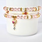 Three stacked bracelets with pink beads, pearls, and gold accents. The top and bottom bracelets have clear, faceted beads. The middle bracelet is made of small pearls. A heart-shaped charm with a floral design and a small crystal dangle hangs from the bottom bracelet.