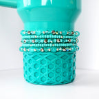 A gumdrop green Stanley with a textured geometric pattern on the bottom. Several beaded bracelets in shades of turquoise and silver are stacked around the middle of the tumbler. The bracelets feature various bead sizes and textures, creating a layered effect.