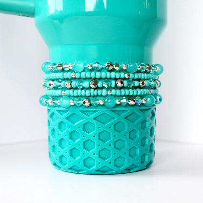 A gumdrop green Stanley with a textured geometric pattern on the bottom. Several beaded bracelets in shades of turquoise and silver are stacked around the middle of the tumbler. The bracelets feature various bead sizes and textures, creating a layered effect.