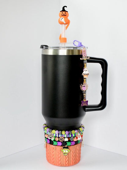 Black tumbler with a handle, adorned with Halloween-themed charms and a decorative sleeve. The sleeve is orange with a cable knit design. A series of beaded bracelets in purple, green, black, and orange encircle the base. A pumpkin-shaped straw topper sits on top, adding a playful touch.